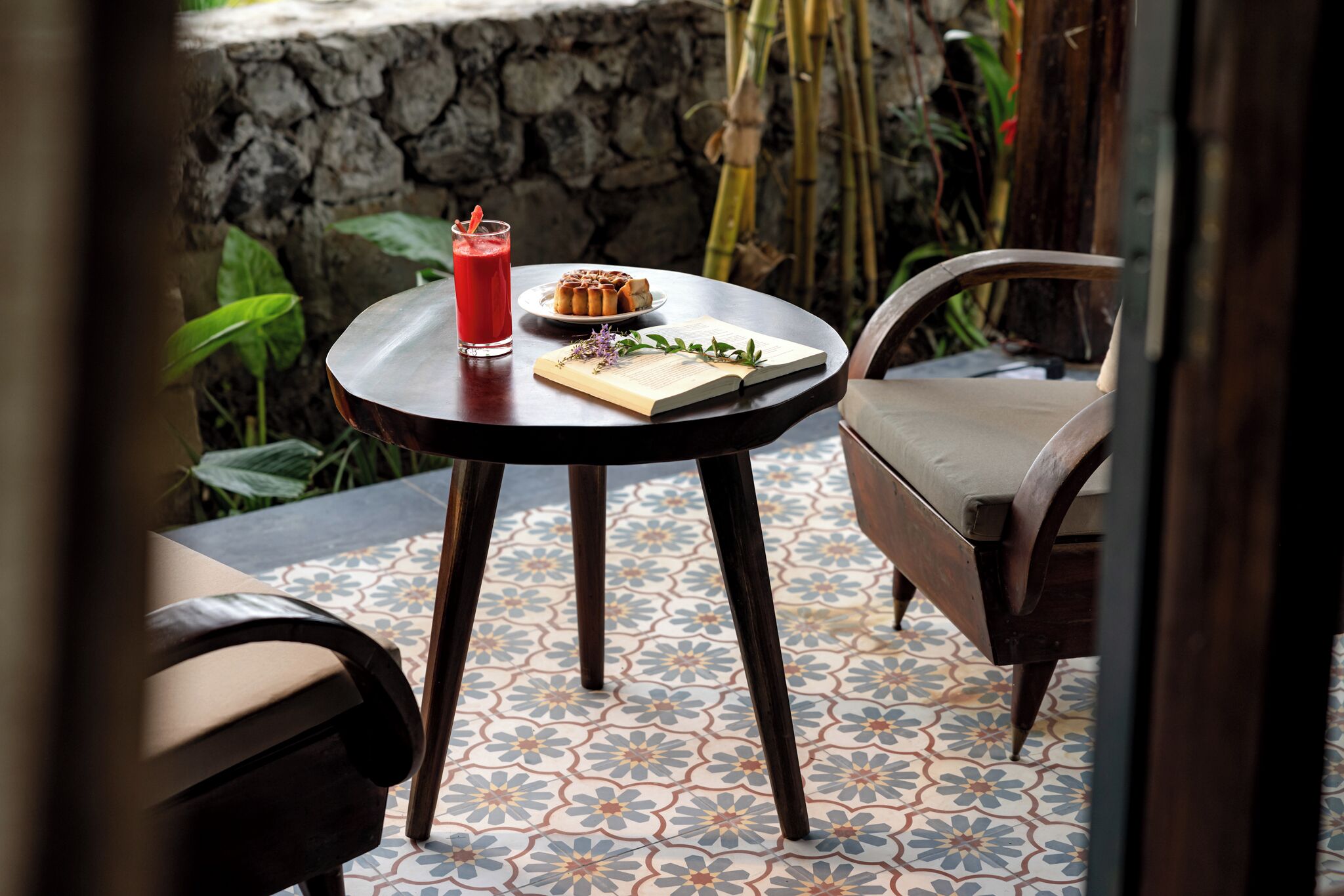 A round table with a book, a plate with food, and a drink on a patterned floor with a stone wall and plants in the background.