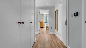 Un pasillo con piso de madera, paredes blancas y puertas que dan a una habitación con cama y ventana.