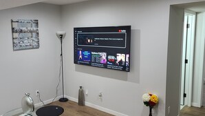 Een moderne woonkamer met een tv aan de muur waarop verschillende programma's te zien zijn, een vloerlamp, een vaas met bloemen en een decoratief object op de vloer.