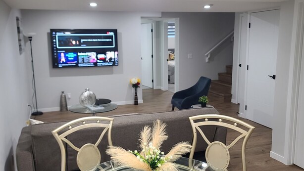 Een moderne woonkamer met een flatscreen televisie aan de muur, een bank, een eettafel met stoelen en een trap naar een andere kamer.