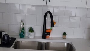 Een keuken met witte kastjes, een roestvrijstalen spoelbak en een zwarte kraan. Op het aanrecht staan potplanten en een fles afwasmiddel.