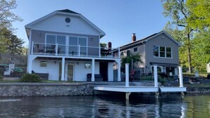 A two-story house with a dock extending into a body of water, surrounded by greenery.