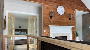 A wooden interior with a fireplace, a clock, and a staircase leading to another room.