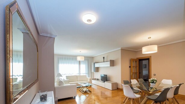 Un salón moderno con un sofá blanco, una mesa de centro de vidrio y una zona de comedor con una mesa y sillas de vidrio.