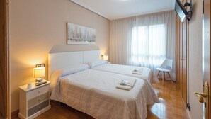 Habitación de hotel con dos camas, una mesita de noche con lámpara, una silla y un cuadro en la pared.