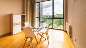 A modern living room with a glass dining table, two chairs, a wooden cabinet, and a large window offering a view of a cityscape and greenery.