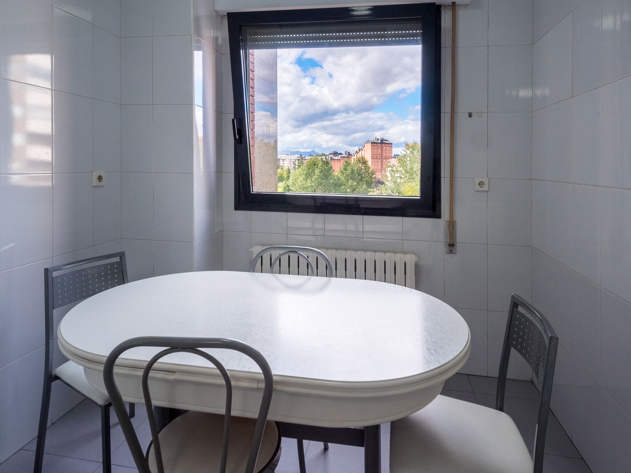 Ein Essbereich mit weißem Tisch und Stühlen, einem Fenster mit Blick auf Gebäude und Bäume sowie einem Heizkörper unter dem Fenster.
