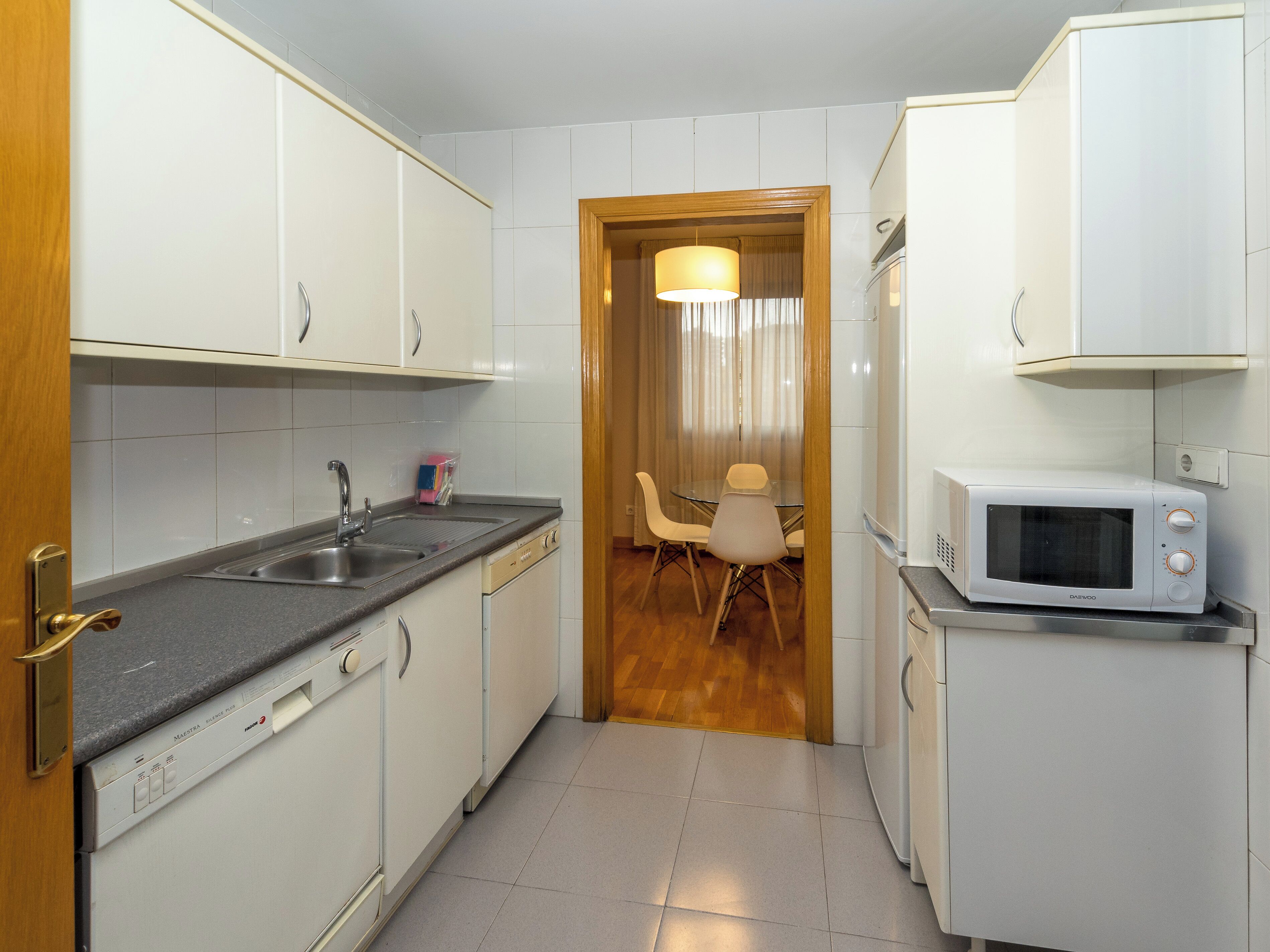 Una cocina compacta con armarios blancos, lavavajillas integrado, microondas y comedor con mesa y sillas.