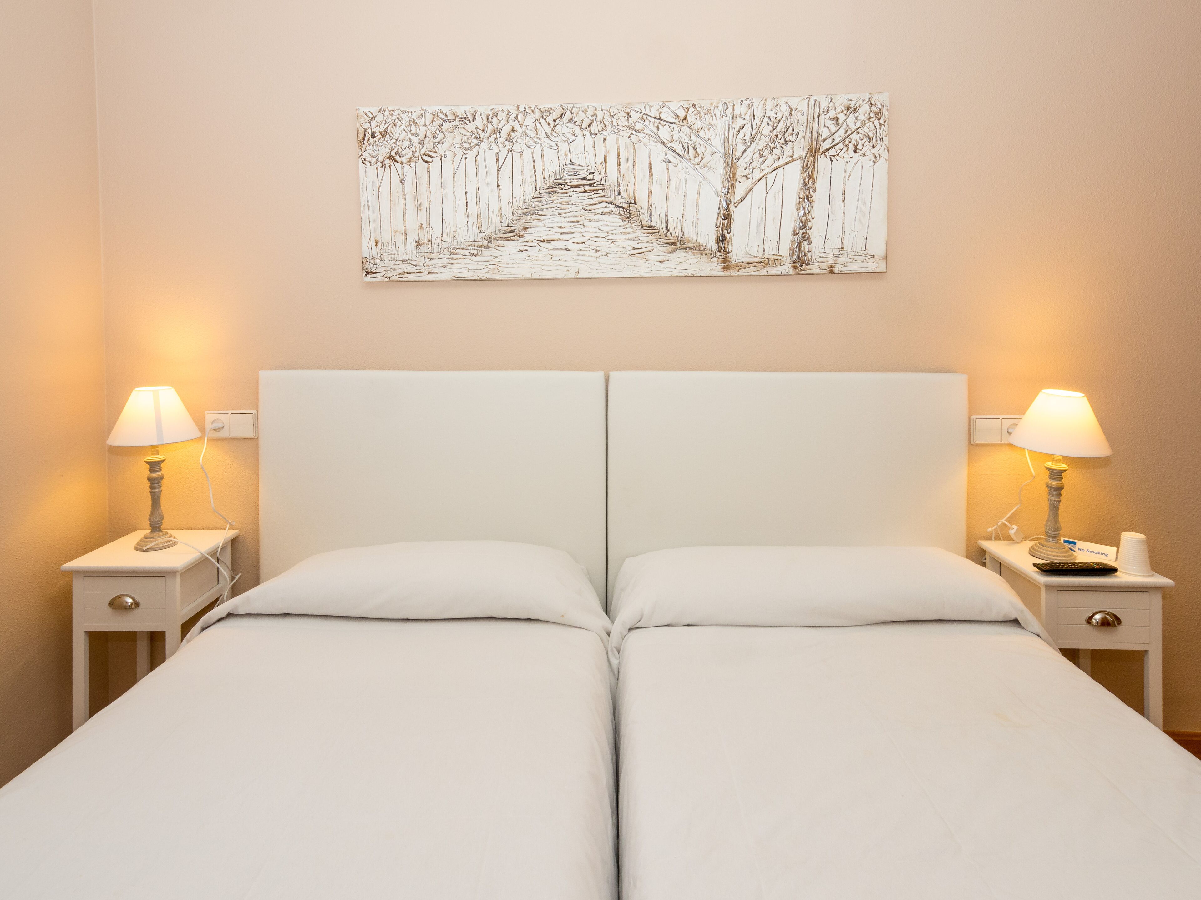 Una cama doble con sábanas blancas, dos mesitas de noche con lámparas y una obra de arte en la pared que muestra un sendero bordeado de árboles.