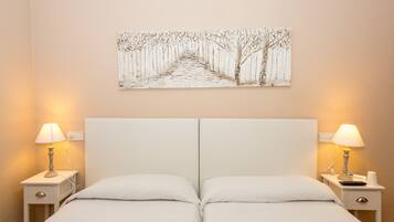 Una cama doble con sábanas blancas, dos mesitas de noche con lámparas y una obra de arte en la pared que muestra un sendero bordeado de árboles.