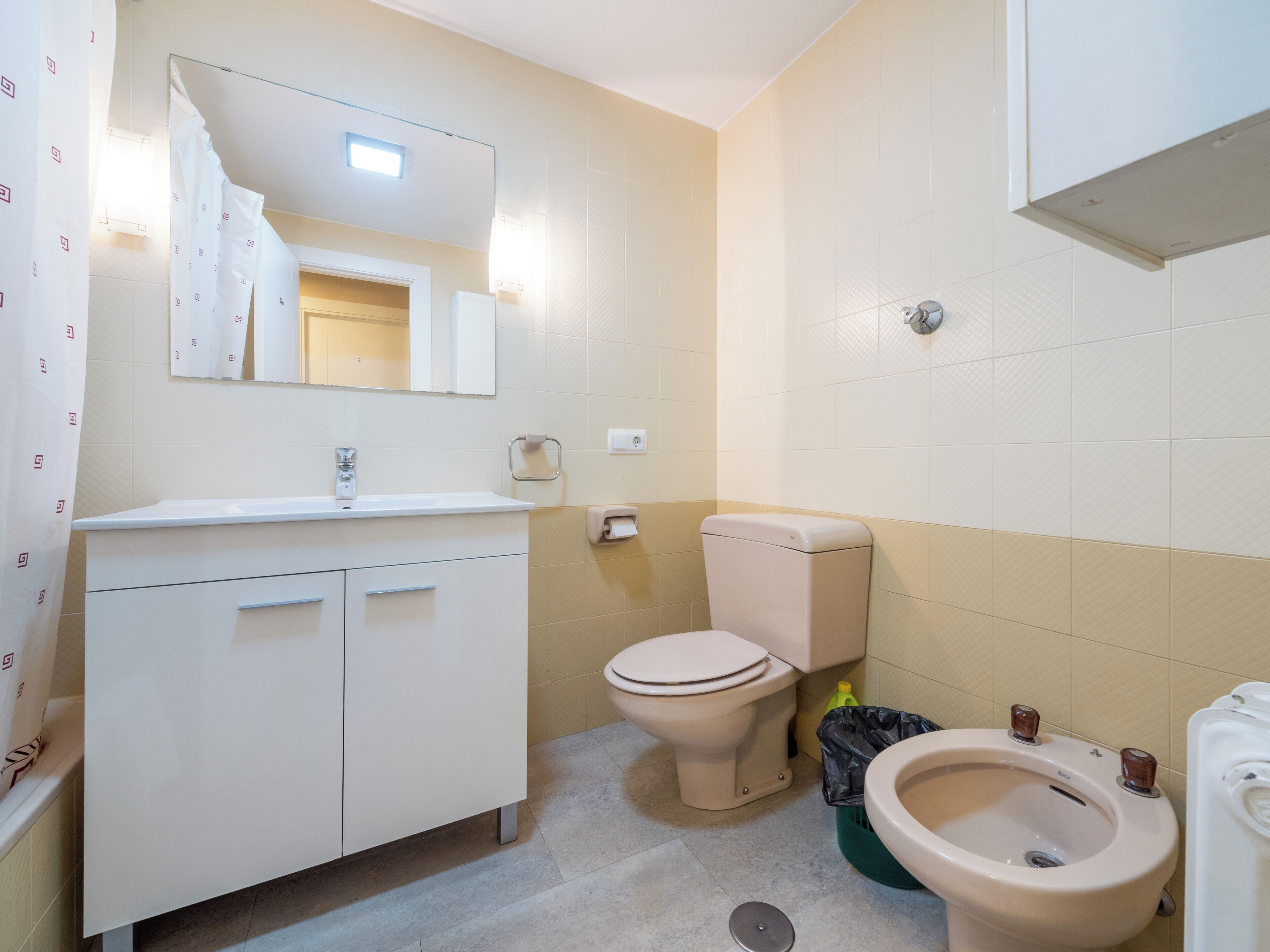 Un baño con lavamanos blanco, espejo, inodoro, bidé y un cubo de basura.