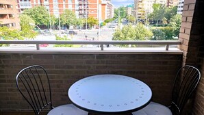 Ein Balkon mit einem runden Tisch und zwei Stühlen, mit Blick auf eine Stadtlandschaft mit Gebäuden und Bäumen.