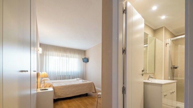 A hotel room with a bed, bedside table with a lamp, a TV, a bathroom with a sink and mirror, and a shower.