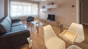A modern living room with a sofa, coffee table, and chairs. There is a TV on a stand and a bookshelf in the background.