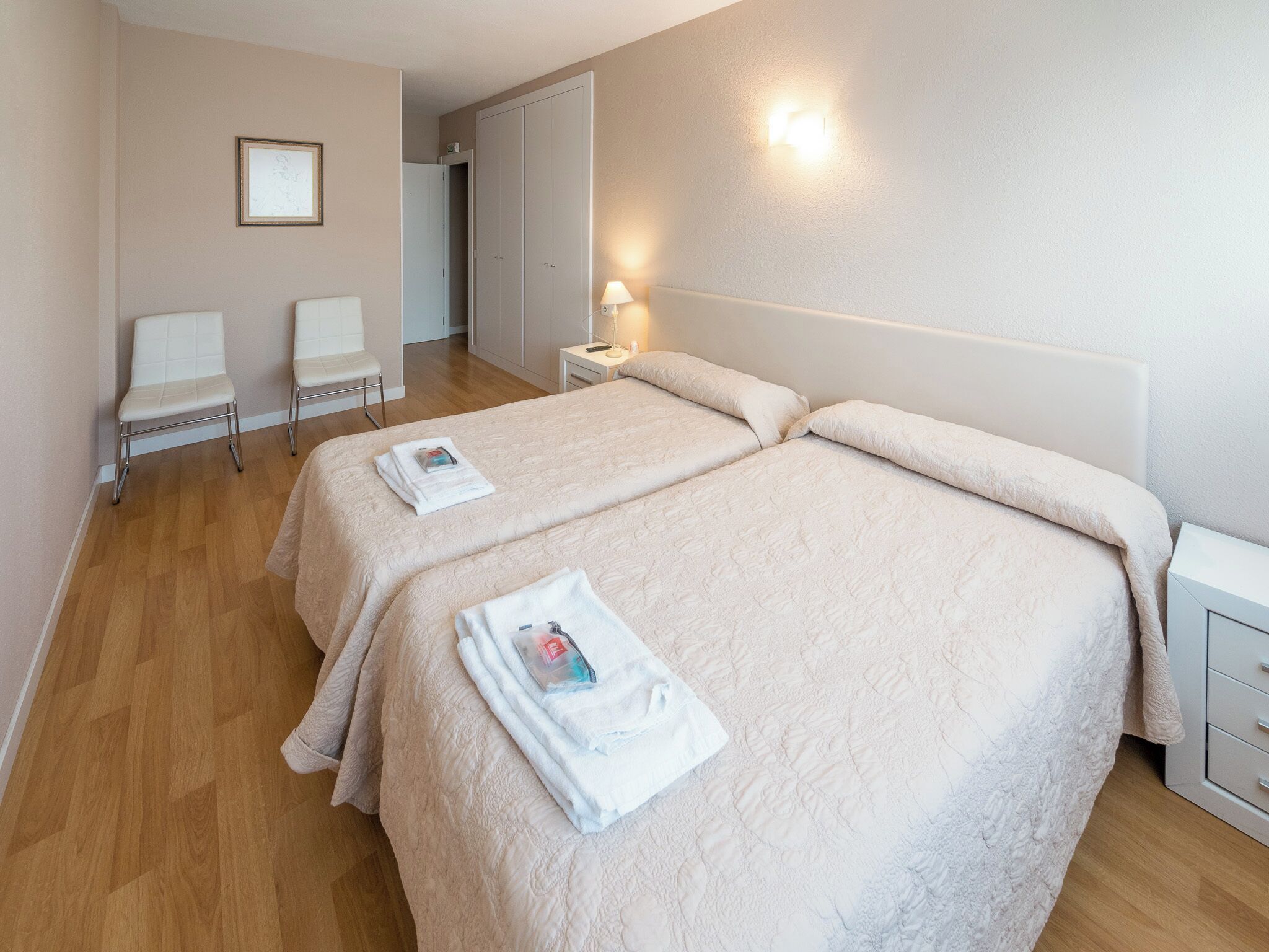 Een hotelkamer met twee bedden, een nachtkastje met een lamp, een stoel en een ingelijste foto aan de muur.