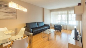 A modern living room with a black leather sofa, a glass coffee table, white chairs, and a television on a wooden stand.