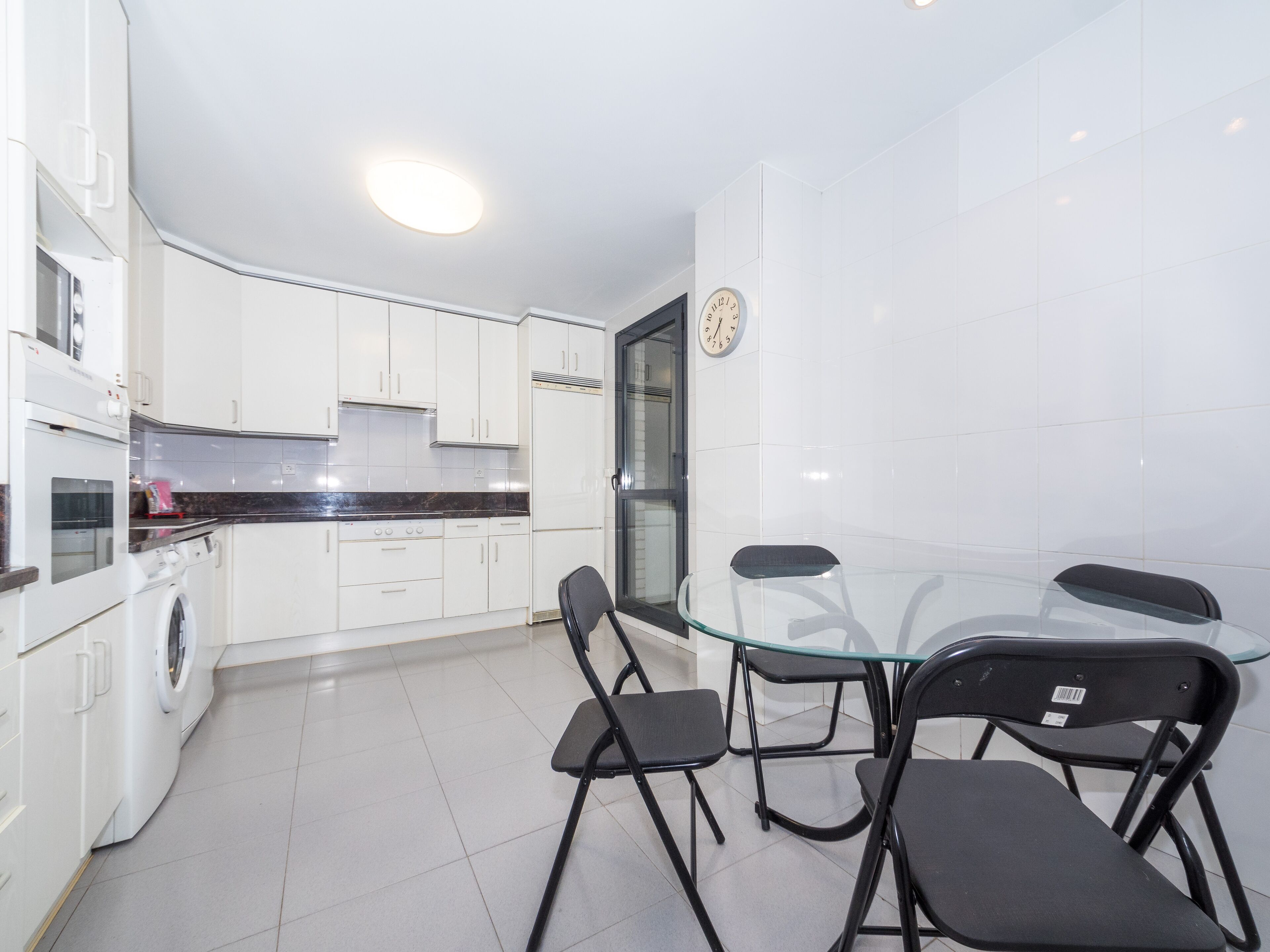 Een moderne keuken met witte kasten, een ingebouwde oven en een glazen eettafel met vier stoelen.