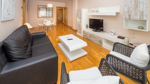 A modern living room with a black leather sofa, white coffee table, wicker chairs, a flat-screen TV, and a dining area with a glass table and chairs. - Apartamento Zona Clínica Universitaria (Pamplona)