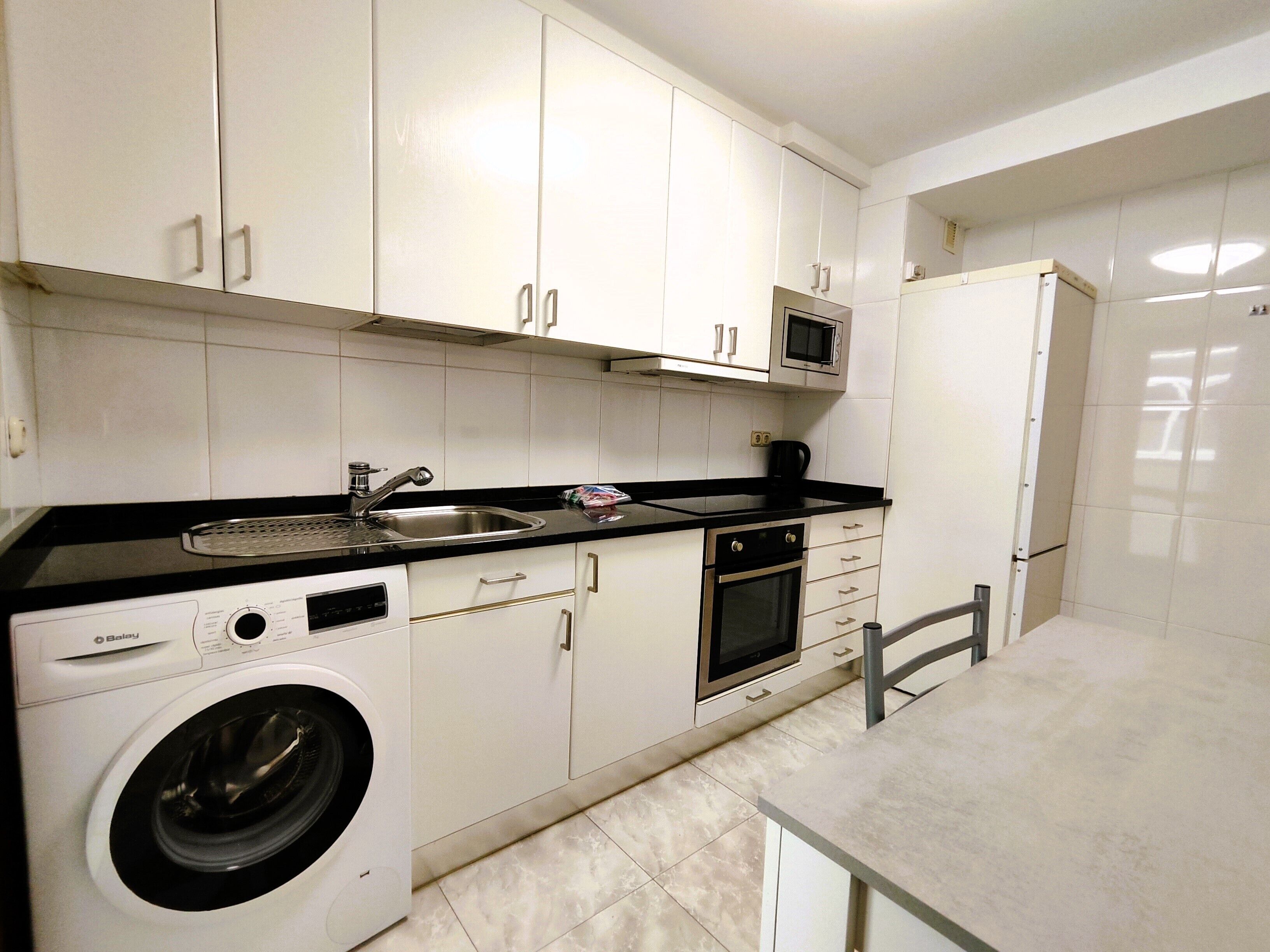Une cuisine moderne avec des meubles blancs, un évier en acier inoxydable, un four à micro-ondes, un four traditionnel et un lave-linge.