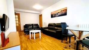 A living room with a wooden floor, two black leather sofas, a white coffee table, a glass table with chairs, a painting on the wall, and a TV on the wall. - Para 4, el Apartamento Idóneo (Pamplona)