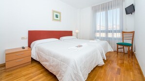 A hotel room with a wooden floor, a red headboard, two beds with white bedding, a small bedside table, a chair, a TV mounted on the wall, and a window with sheer curtains. - Para 4, el Apartamento Idóneo (Pamplona)