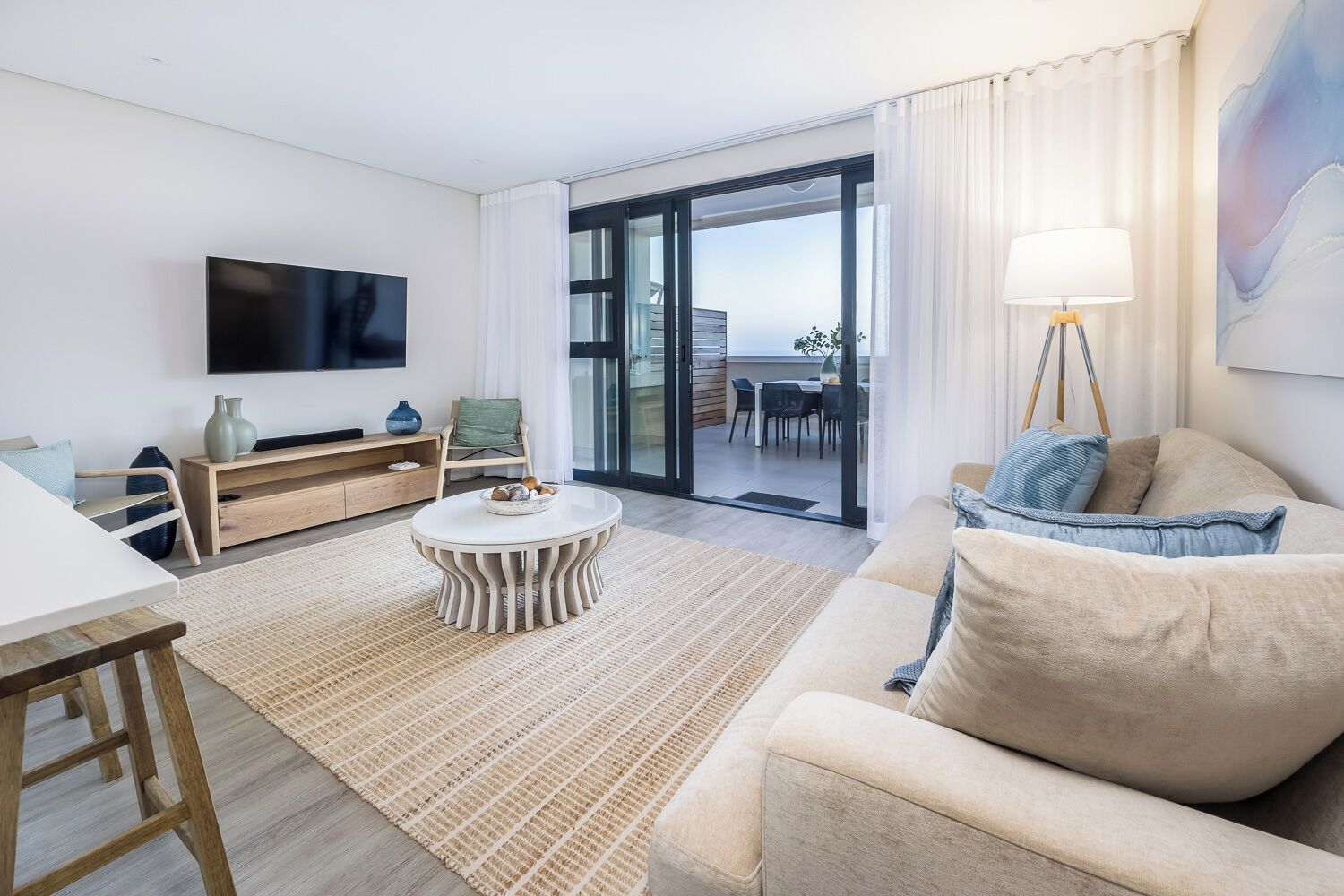 A modern living room with a flat-screen TV, a round coffee table, a sofa, and a dining area visible through a glass door.