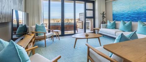 A modern living room with a large sliding glass door leading to a balcony, a flat-screen TV, and a painting of the ocean.