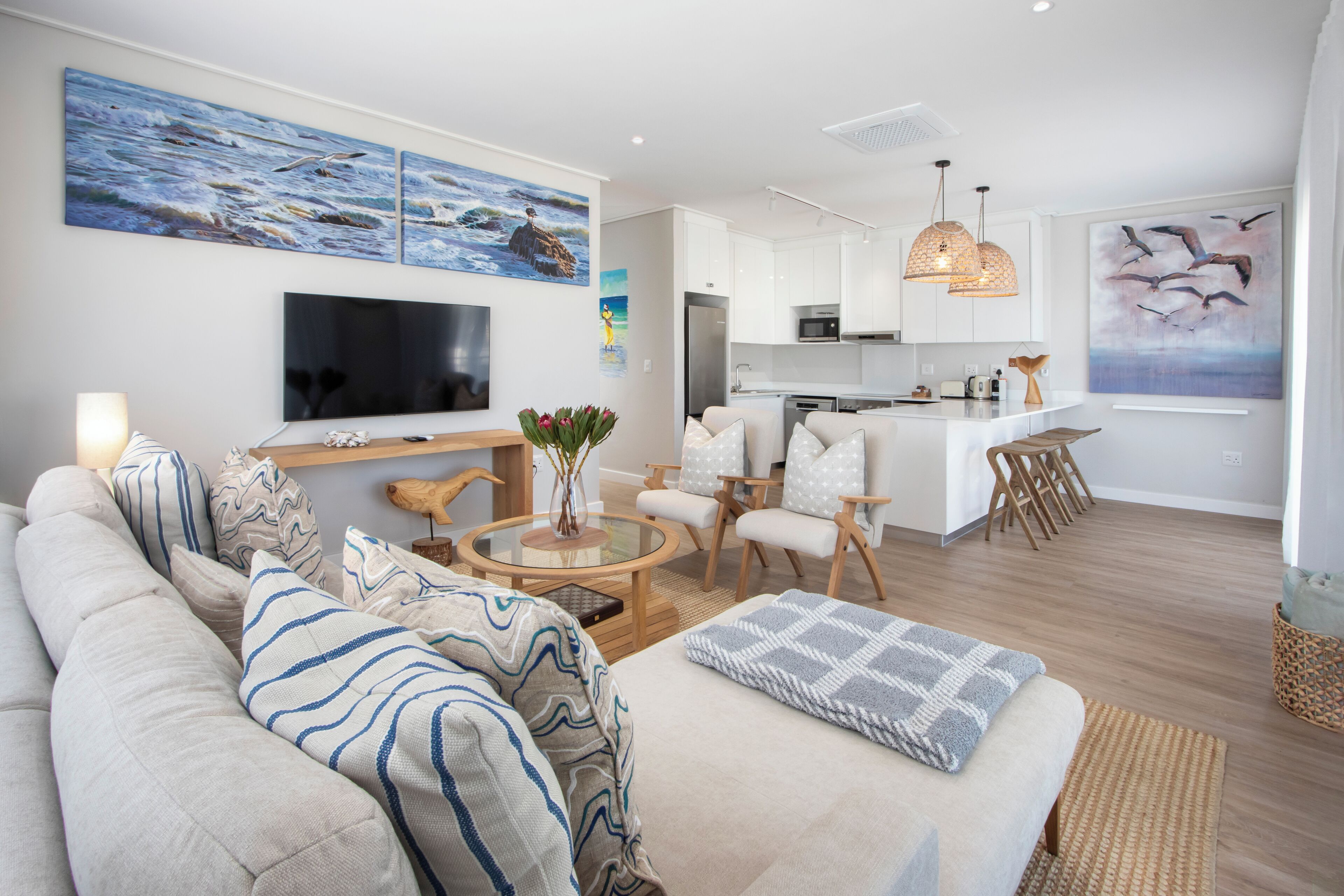 A modern living room with a sofa, coffee table, and TV. There is a kitchen area with white cabinets and a dining space with chairs.