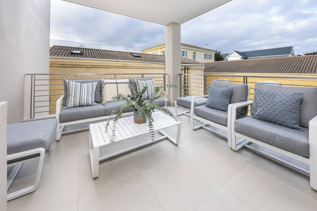 Un espace extérieur moderne, aménagé avec des meubles blancs, une table basse centrale et offrant une vue sur des immeubles d’habitation.