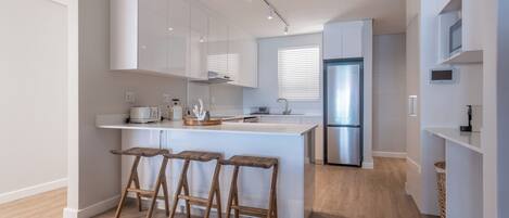 Una cocina moderna con armarios blancos, una isla central con taburetes, un refrigerador de acero inoxidable y una ventana con persianas.