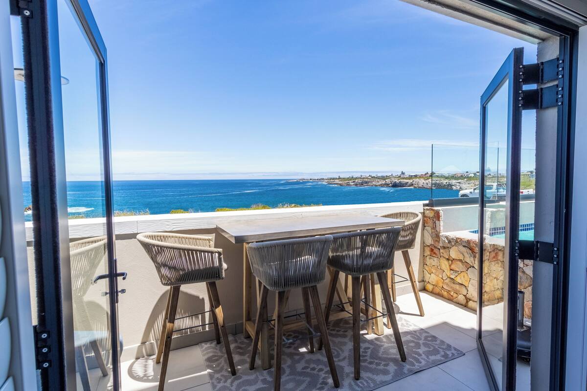 Une porte coulissante s’ouvrant sur un balcon aménagé avec une table et des chaises, d’où l’on peut admirer une vue sur la côte.