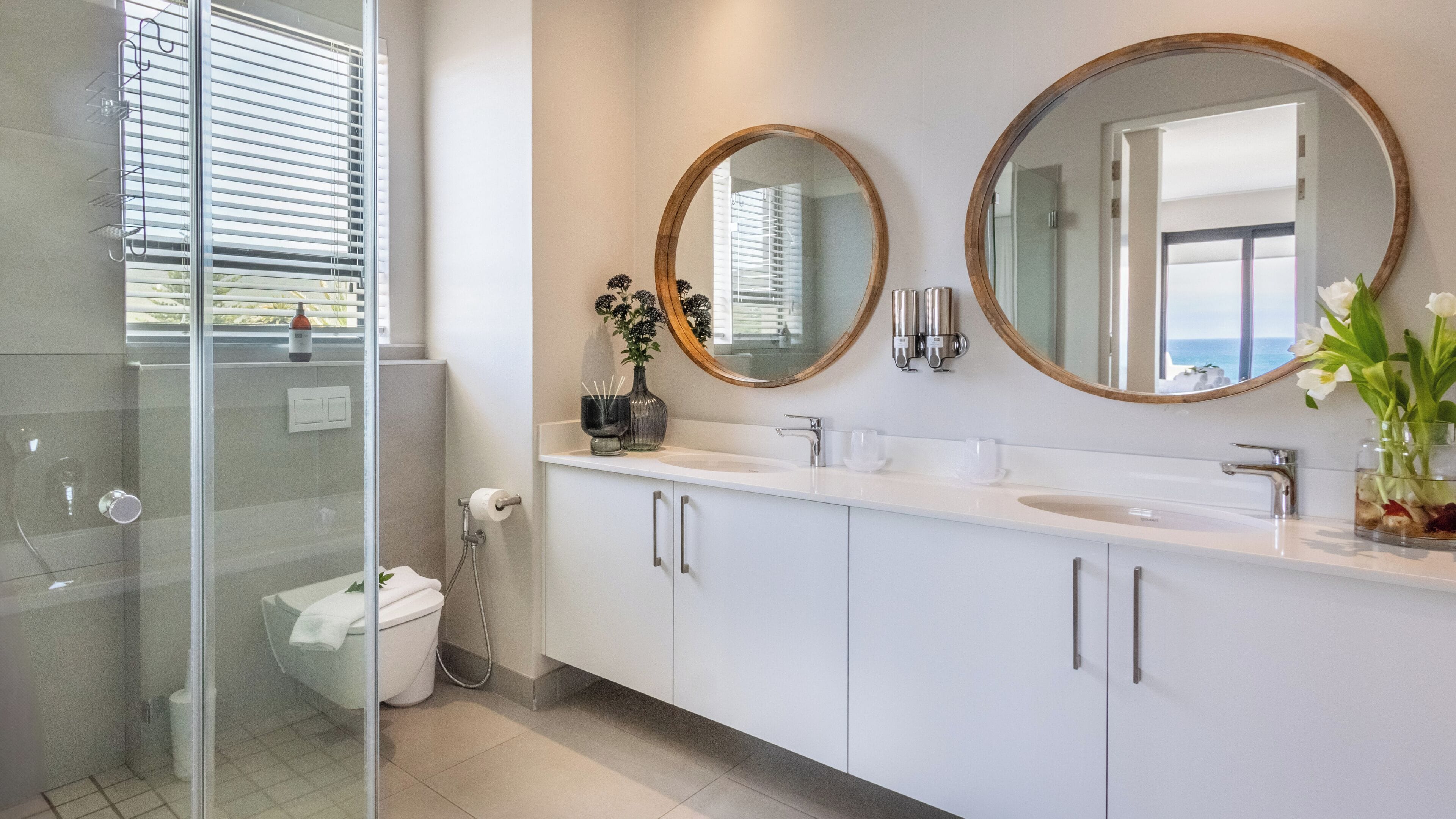 A modern bathroom with a glass shower enclosure, a white sink with two round mirrors, and a vase with flowers on the countertop.
