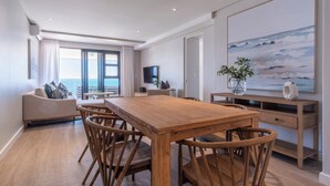 A modern living room with a wooden dining table, wicker chairs, a sofa, and a large painting.