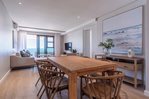 A modern living room with a wooden dining table, wicker chairs, a sofa, and a large painting.
