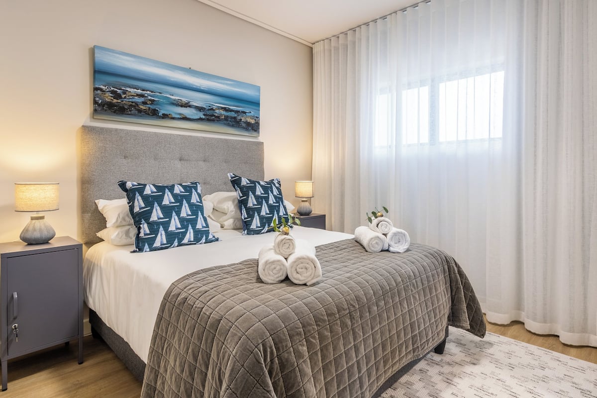 Une chambre d’hôtel bien rangée, avec un lit, deux tables de chevet, un tableau et une fenêtre avec des rideaux.