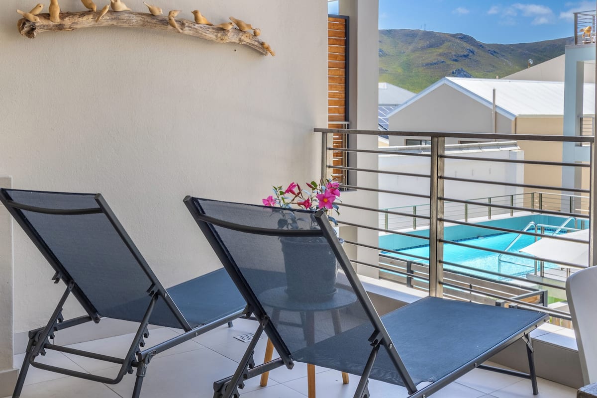 Un balcon avec deux chaises en treillis noir, une petite table et une branche décorative en bois ornée de figurines d’oiseaux. Un vase rempli de fleurs roses est posé sur la table. On aperçoit une piscine à travers la rambarde du balcon.