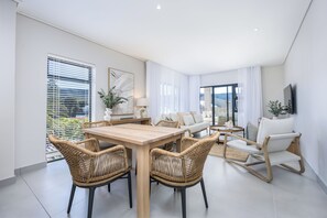 A modern living room with a wooden dining table, wicker chairs, a white sofa, and a TV mounted on the wall.