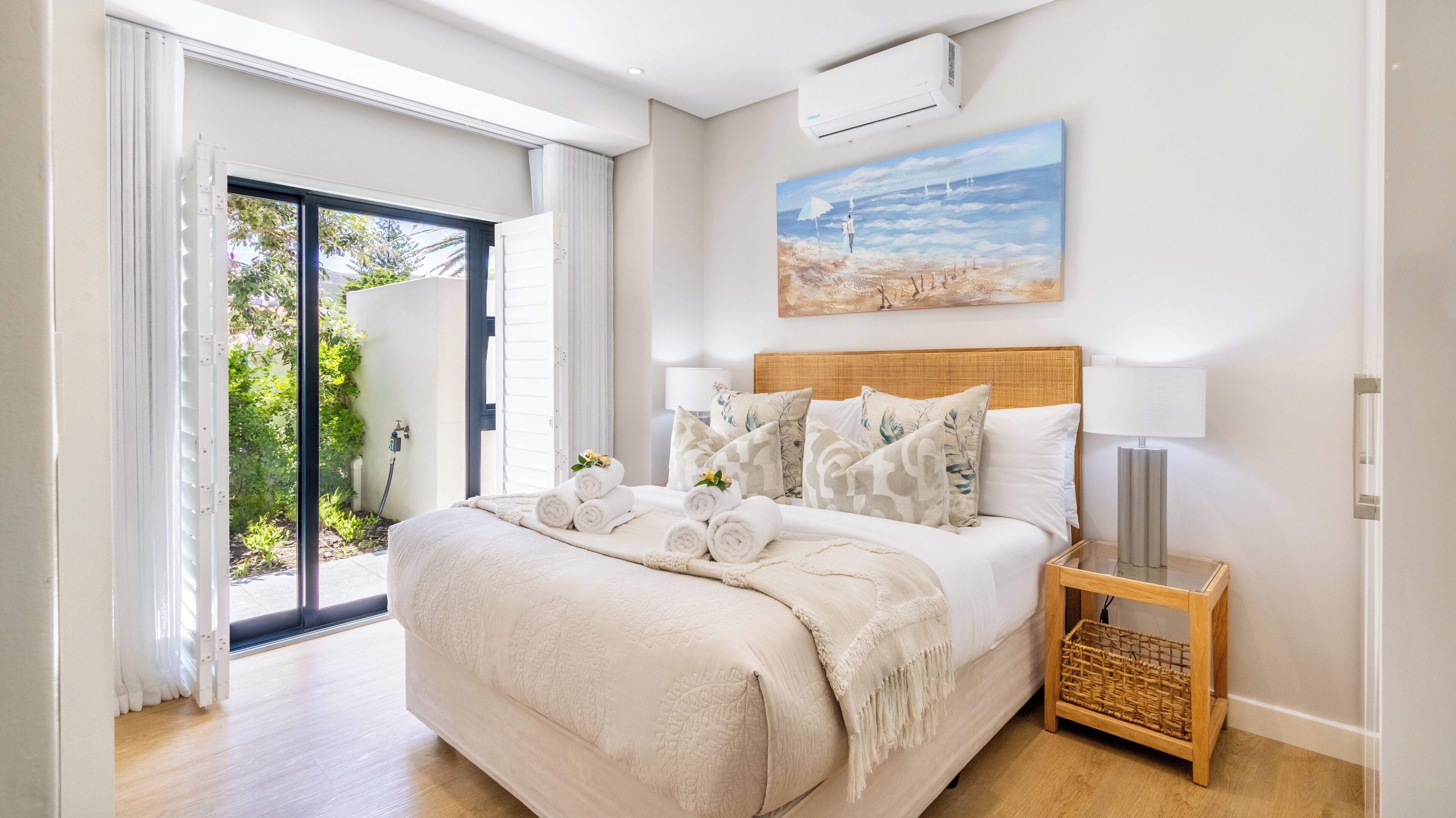 Een slaapkamer met een groot bed, een schilderij van een strandgezicht, een klein nachtkastje en uitzicht op groen buiten.