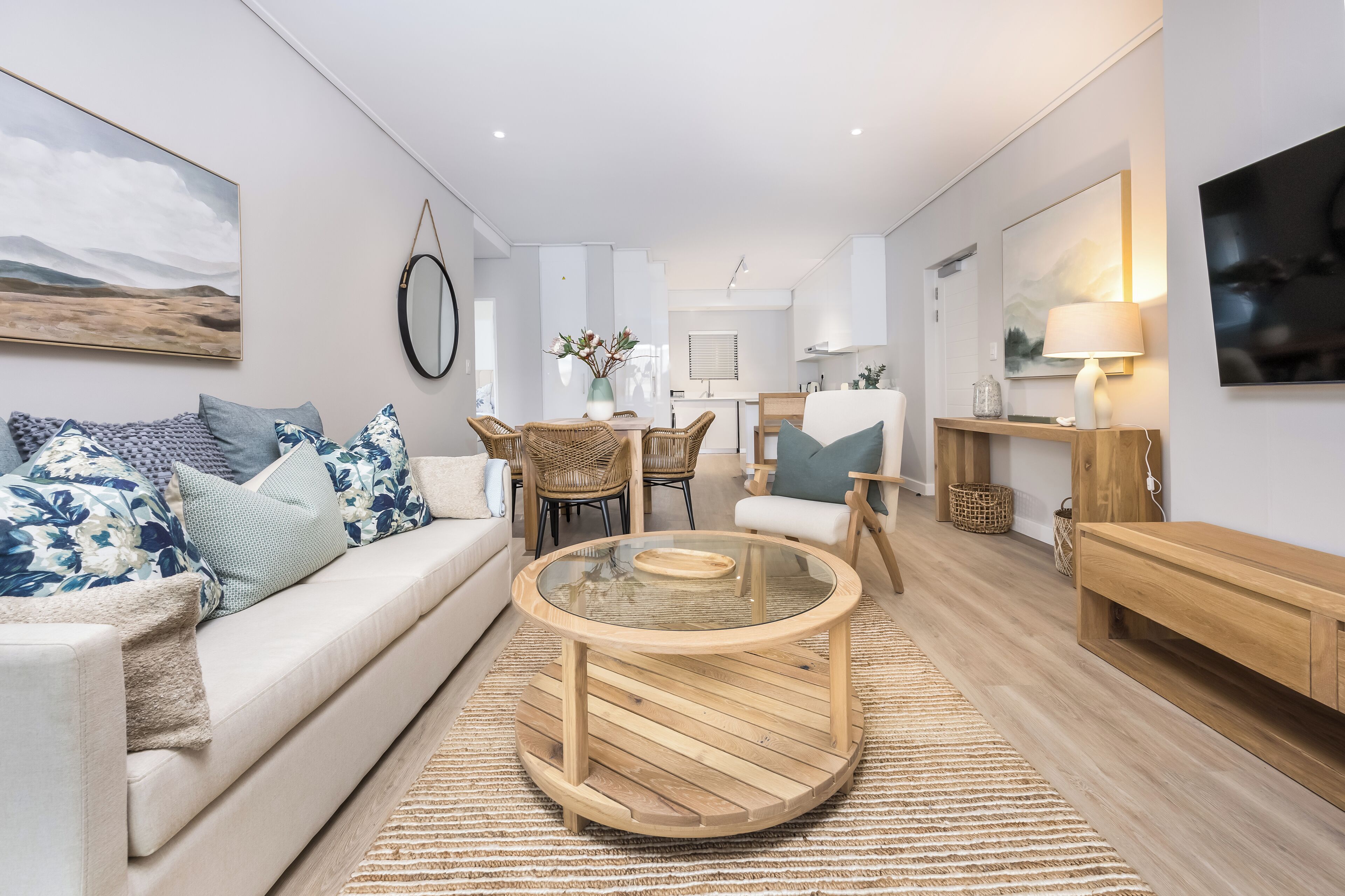 A modern living room with a round wooden coffee table, a sofa with patterned cushions, a dining area with wicker chairs, and a kitchen in the background.