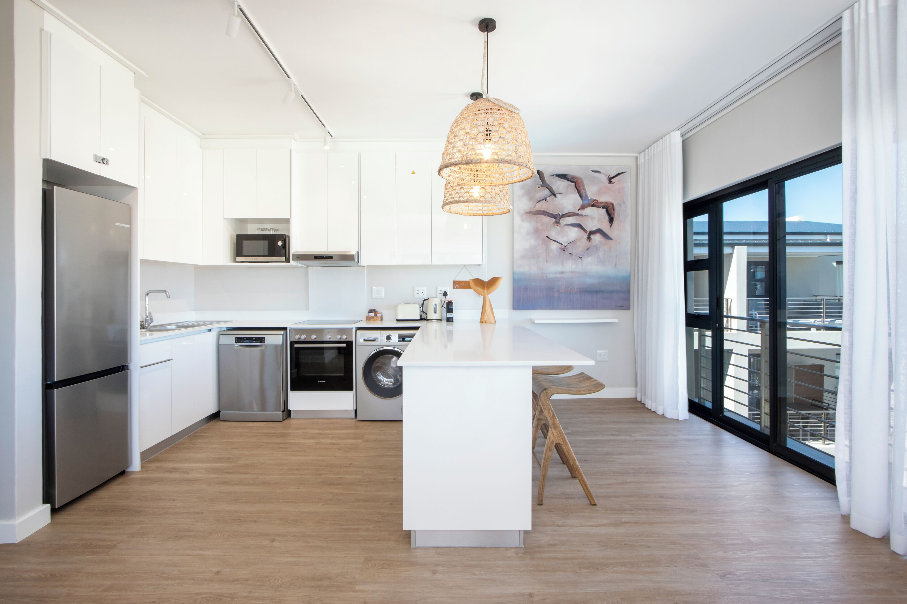 Cocina moderna con armarios blancos, electrodomésticos de acero inoxidable, una isla central y un amplio ventanal con vistas.