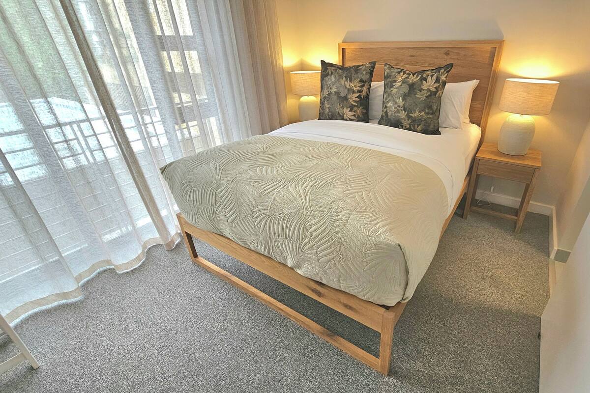 Un lit bien fait, recouvert d’une couvre-lit à motifs, avec deux oreillers et une tête de lit en bois. On y trouve une table de chevet avec une lampe et une petite étagère. La pièce est dotée d’un sol recouvert d’une moquette et d’une fenêtre ornée de rideaux légers.