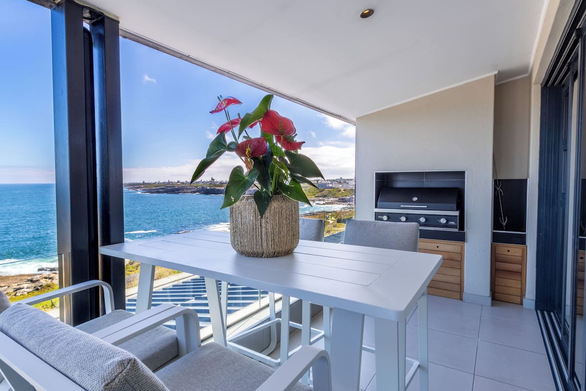 Un balcon avec une table et des chaises blanches, une plante en pot et un barbecue.