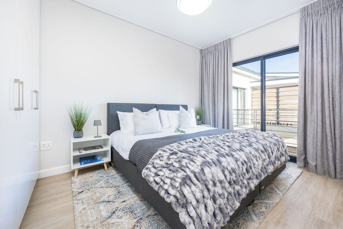 Une chambre moderne avec un grand lit, une table de chevet, une plante en pot et une vue sur l’extérieur grâce à une grande fenêtre.