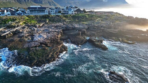 A coastal hotel with a rocky shoreline, ocean waves, and a mountainous backdrop.