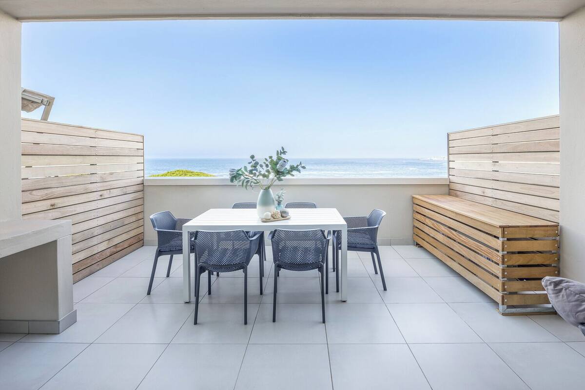 Une terrasse avec une table et des chaises blanches, un vase rempli de fleurs et une vue sur la mer.