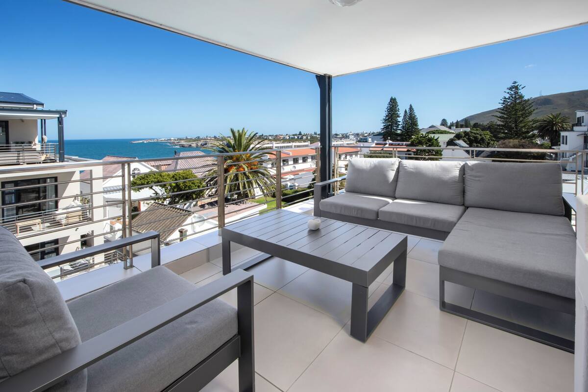 Un balcon moderne doté d’un canapé, d’une table et de chaises, offrant une vue sur la mer et les bâtiments environnants.