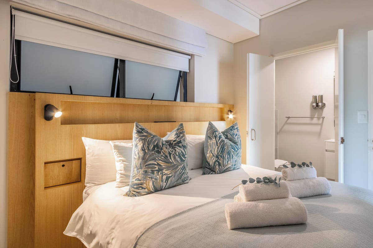 Une chambre à coucher moderne avec une tête de lit en bois, un grand lit agrémenté d’oreillers à motifs, une table de chevet et une vue sur une salle de bain équipée d’un porte-serviettes.