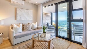Un salón moderno con sofá, una mesa de centro de vidrio y vista al océano a través de amplios ventanales correderas.