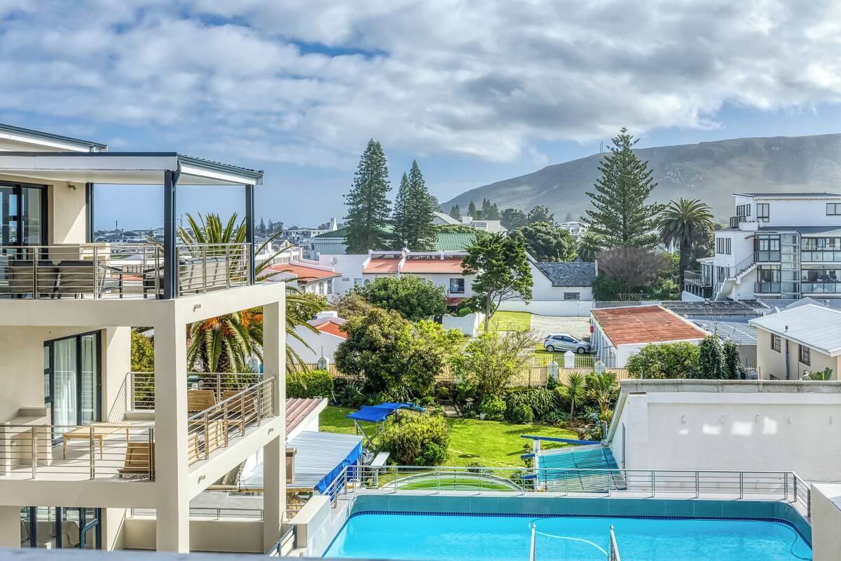 Un bâtiment moderne doté d’une piscine, d’un espace extérieur couvert et offrant une vue sur une montagne et les maisons environnantes.