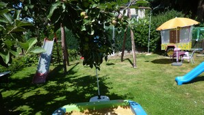 Un arenero con borde azul, un árbol con hojas verdes y manzanas, y una estructura de sombrilla amarilla al fondo.